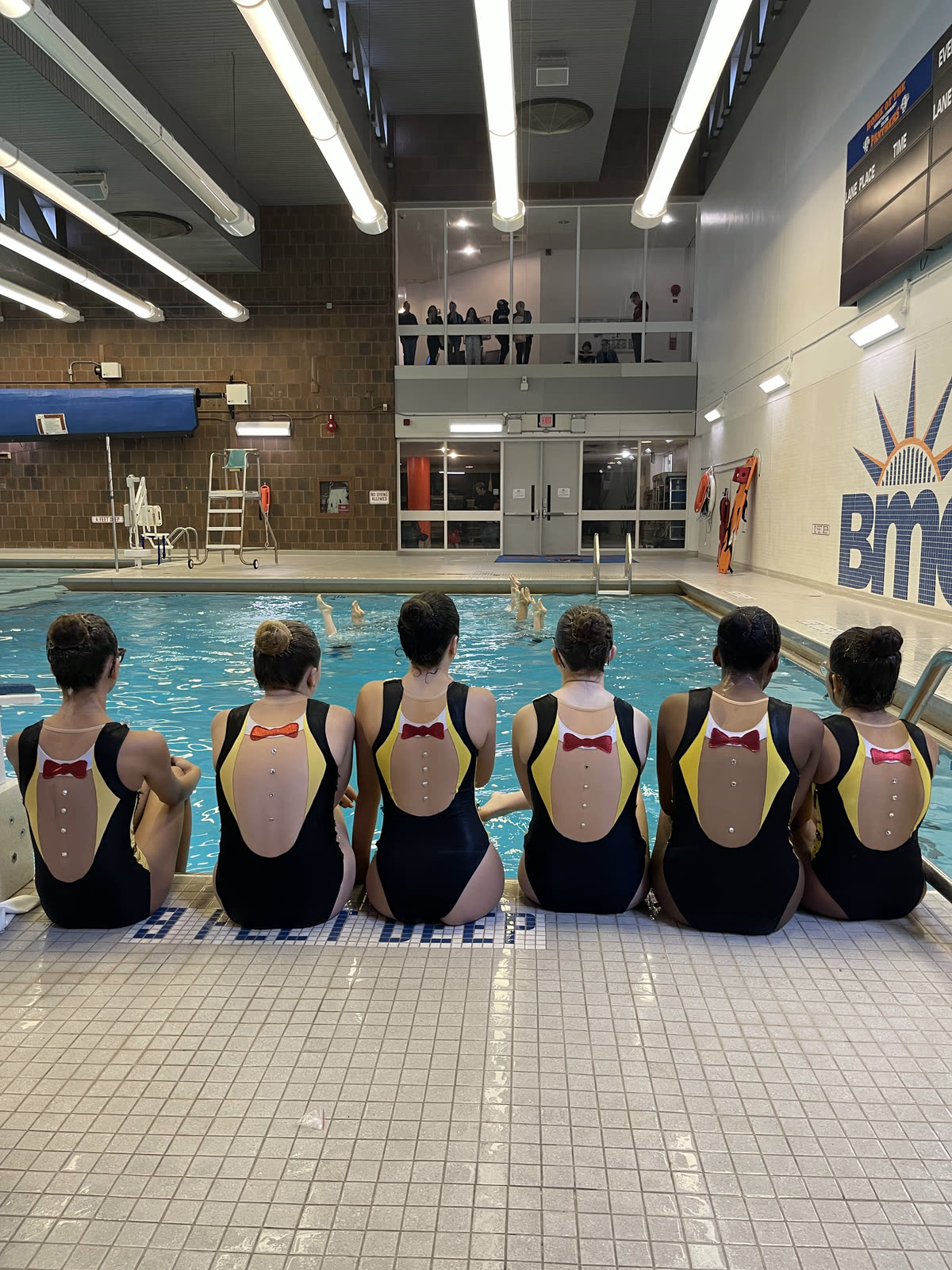 Six swimmers in black and yellow competition suits sitting on the BMCC pool deck, watching teammates rehearse in the water