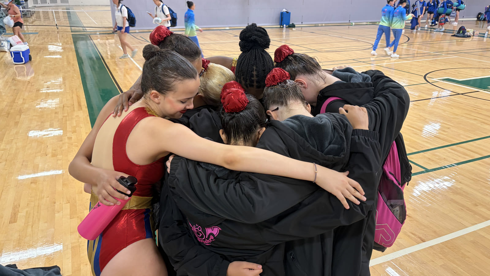 The team in a huddle on a gym floor at the USA Junior Olympics, arms around each other over their competition suits